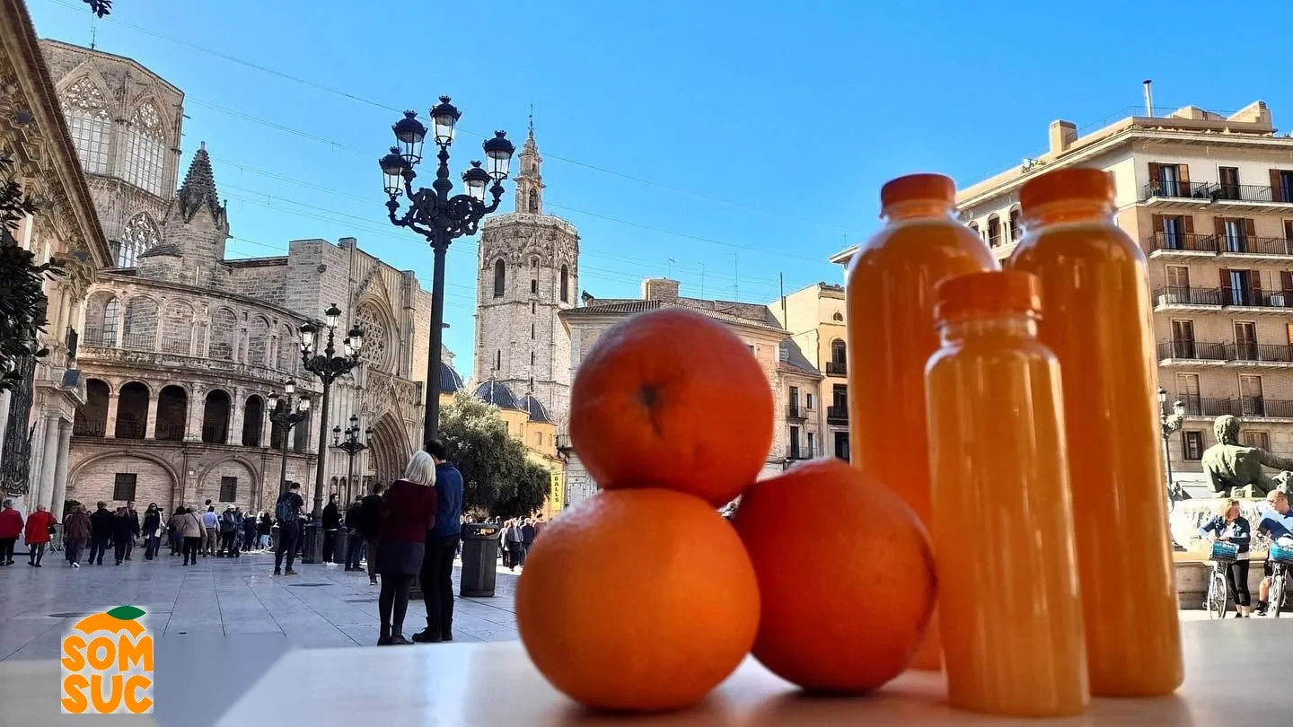 Botellas de zumo de naranja recién exprimido y naranjas en la plaza de la Virgen de València, con el Micalet al fondo