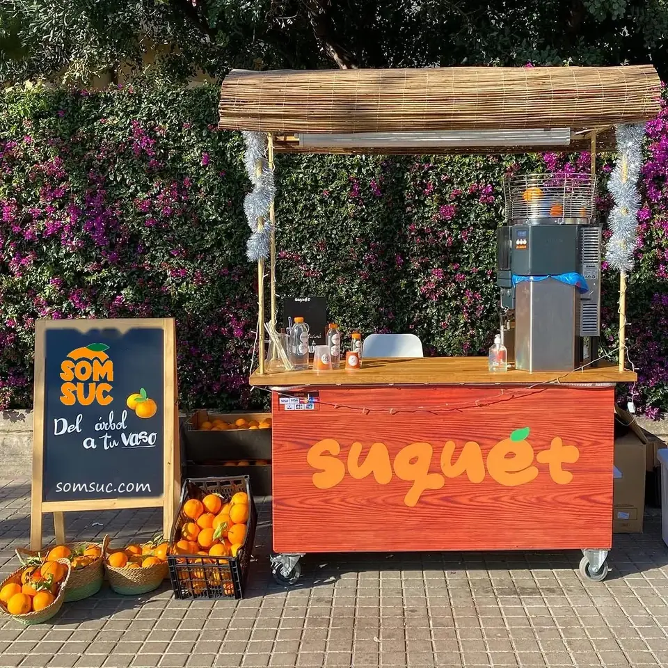 Carrito de zumo de naranja recién exprimido de SOMSUC en València, con cajas y capazos de naranjas de nuestra finca